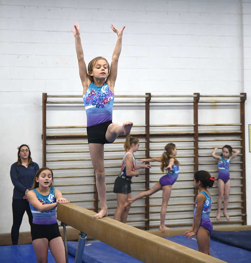 Child performing on a balance beam while their peers and coach look on.